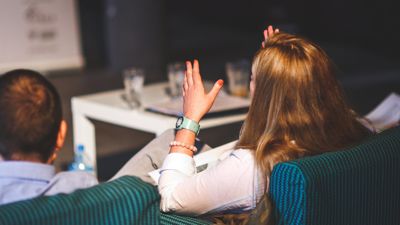 A woman holding her hands up while sitting down at an event - Image taken from behind